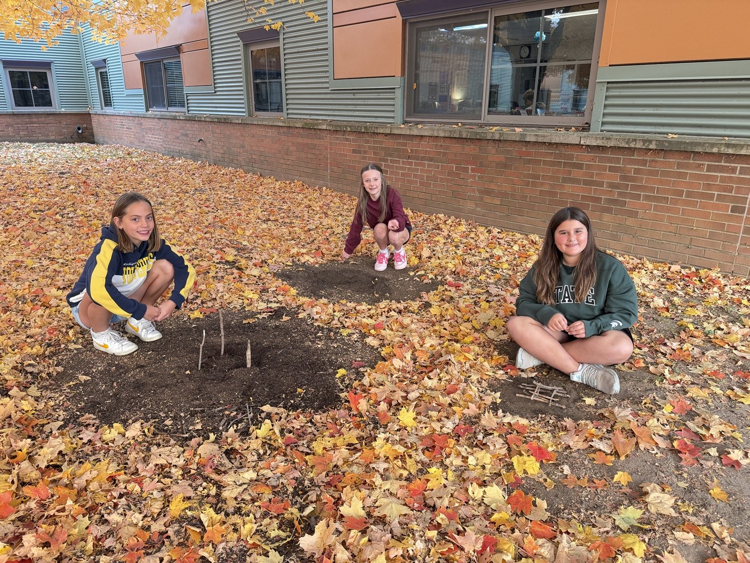 Three students sitting among fallen leaves beside arrangements of sticks.
